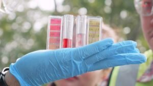 A person wearing blue latex gloves is holding a strip of pH test paper and three test tubes with various levels of red liquid against a background of blurred greenery, indicating a water quality testing process.