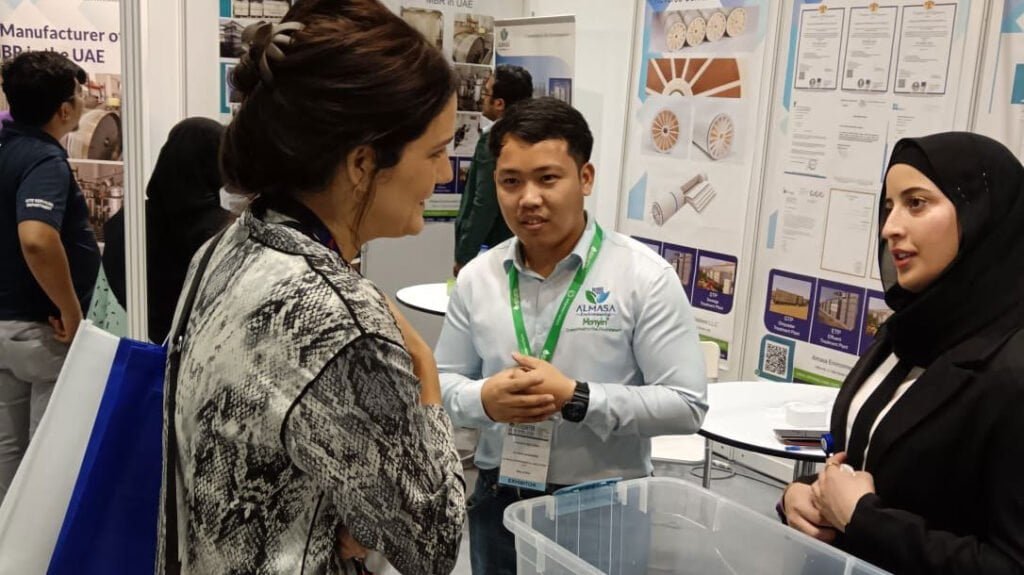 Three people engaged in a conversation at a trade show booth. One man in an 'ALMASA' shirt and two women, one in a hijab, discuss products with posters and brochures displayed in the background.