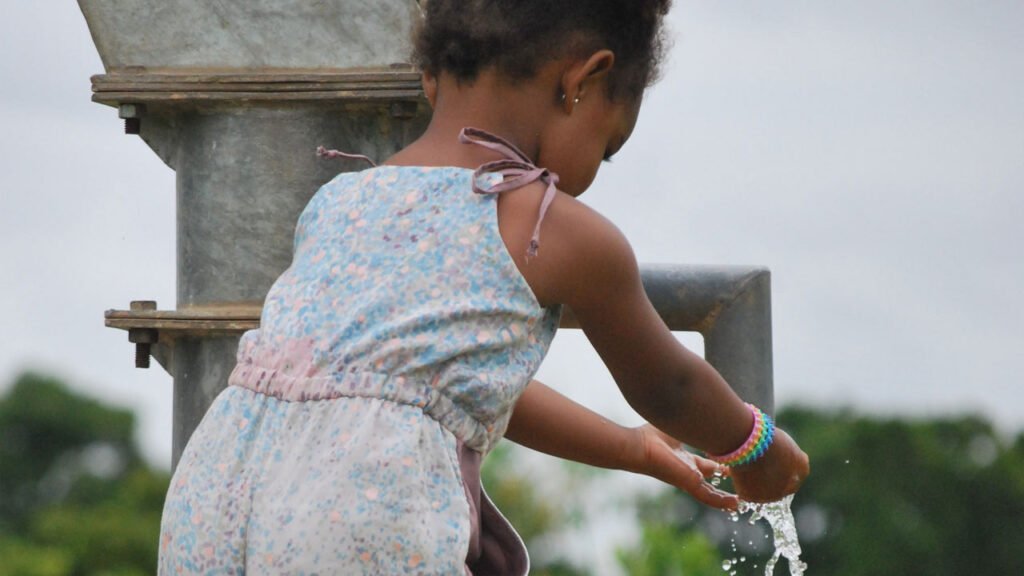 A young girl with curly hair in a colorful jumpsuit and blue shoes washes her hands at a water well. She stands on a concrete platform with green vegetation in the background under a cloudy sky.