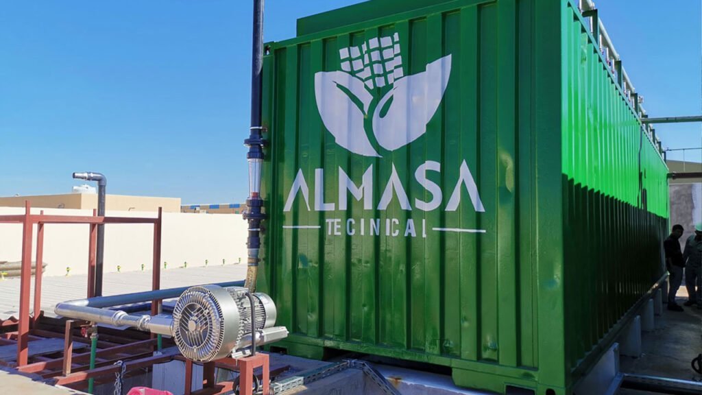 A large green industrial container labeled 'ALMASA TECNICAL' at a water treatment plant. Nearby are metal pipes and a motorized pump. The setup is outdoors under a clear blue sky.