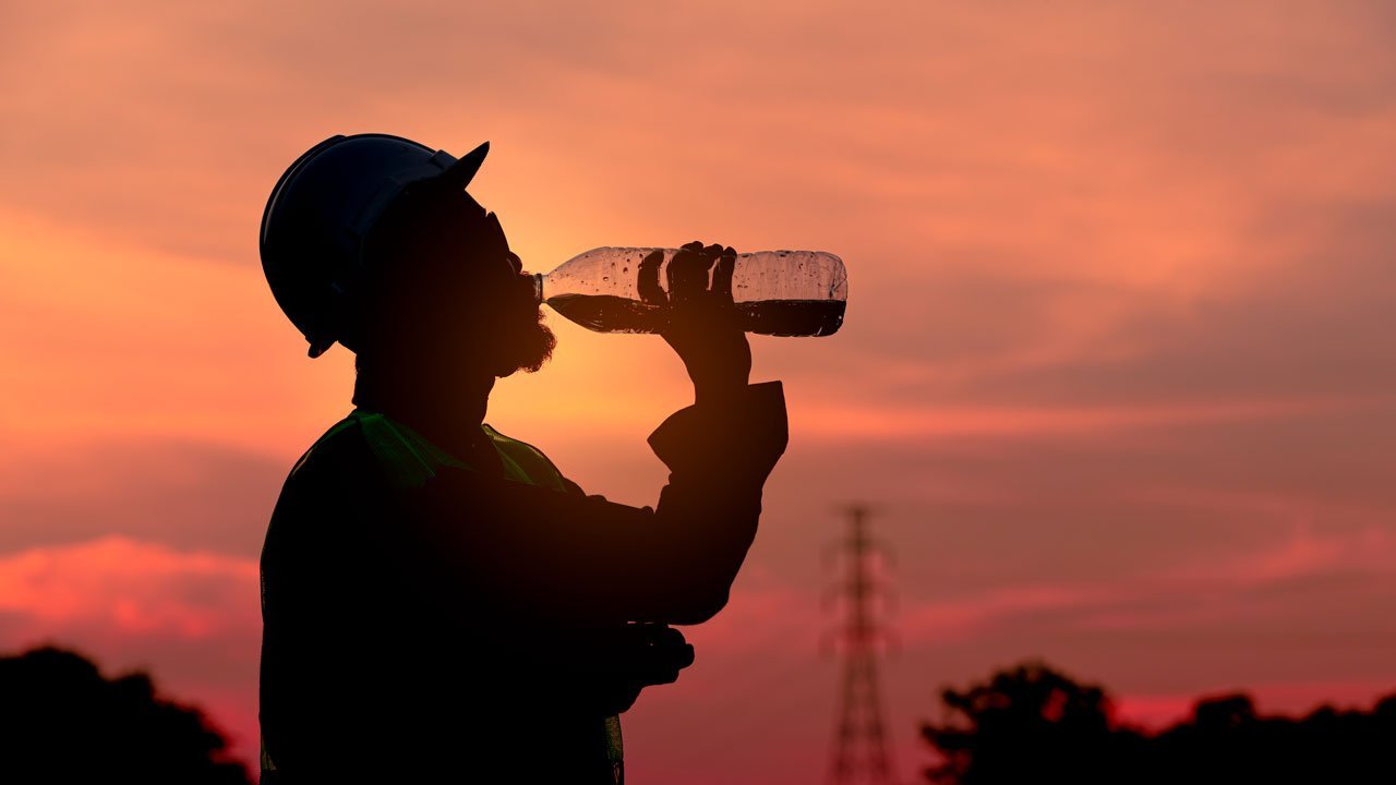 "A silhouette of a worker wearing a hard hat and safety vest drinks from a water bottle at sunset. The sky is a gradient of orange and pink, with trees and an electrical tower in the background.